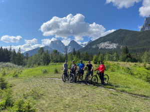 Five male mountain bikers taking a break for a photo during a guided mountain bike ride with Flow Mountain Bike Adventures. Very green landscape with the majestic Canadian Rocky Mountains in the background. The photo was taken on the Loki's mountain bike trail in Canmore, Alberta, Canada. 